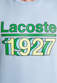Camiseta de algodón azul claro con el logotipo "Lacoste" en verde y "1927" en un diseño ondulado con colores amarillo, azul claro y verde.