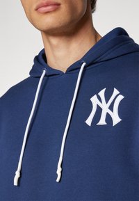 Felpa blu navy con lacci bianchi e logo ricamato bianco sul petto. Il materiale sembra morbido con una vestibilità standard.