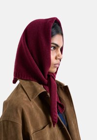 Femme portant un manteau en daim marron et un foulard en tricot bordeaux noué sous le menton, tournant vers la droite sur un fond uni.