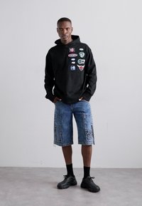 Μαύρη κοντή κουκούλα με διάφορες παρακάμψεις; μπλε τζιν σορτς με πλαϊνές τσέπες; μαύρα ογκώδη αθλητικά παπούτσια; στέκεται σε έναν ανοιχτό γκρίζο πάτωμα.