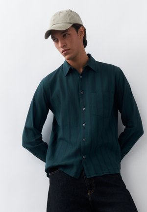 Νέος άνδρας με σκούρο τιρκουάζ πουκάμισο με κουμπιά, μαύρο παντελόνι και μπεζ καπέλο μπέιζμπολ, στέκεται μπροστά σε λευκό φόντο.