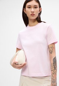 Lyserød bomulds T-shirt med korte ærmer og rund halsudskæring, med et diskret logo. Modellen holder en lille kremfarvet pung.