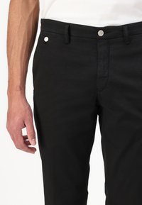Black trousers made from a smooth fabric, featuring a zip fly and button closure, with side pockets and a slim fit design.