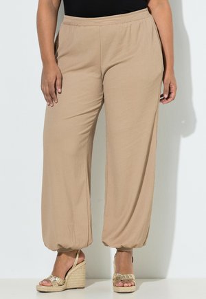 Frau trägt beige, mit elastischem Bund ausgestellte Hosen mit gerafften Säumen und goldenen Keilsandalen, steht vor einer weißen Wand.