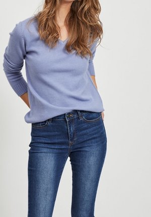Maglione a maglia morbida di un leggero colore viola con scollatura a V e polsini a coste, abbinato a jeans in denim blu scuro, offrendo una silhouette aderente.