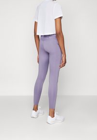 Kobieta ubrana w lila legginsy z wysokim stanem, białą krótką perforowaną bluzkę i białe sneakersy, stojąca na jednolitym tle.