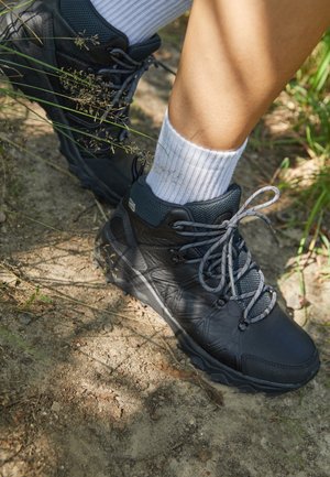 Jambes portant des chaussures de randonnée noires et des chaussettes blanches, debout sur un chemin en terre avec de l'herbe et de petites plantes.