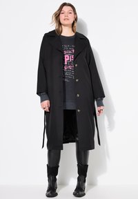 Manteau noir avec un col cranté, design croisé, boutons dorés et ceinture en tissu. Porté par-dessus un sweat-shirt à motif et des leggings.