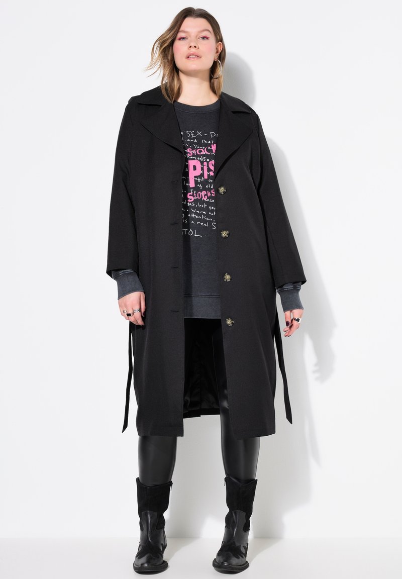 Manteau noir avec un col cranté, design croisé, boutons dorés et ceinture en tissu. Porté par-dessus un sweat-shirt à motif et des leggings.