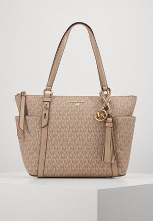 Sac cabas beige Michael Kors avec motif monogramme, doubles poignées, poches latérales, breloque logo dorée et pompon en cuir sur un socle blanc.