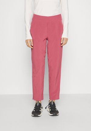 Person iført højtaljede, lige bukser i pink med sidelommer og sorte sneakers, stående mod en ensfarvet baggrund.