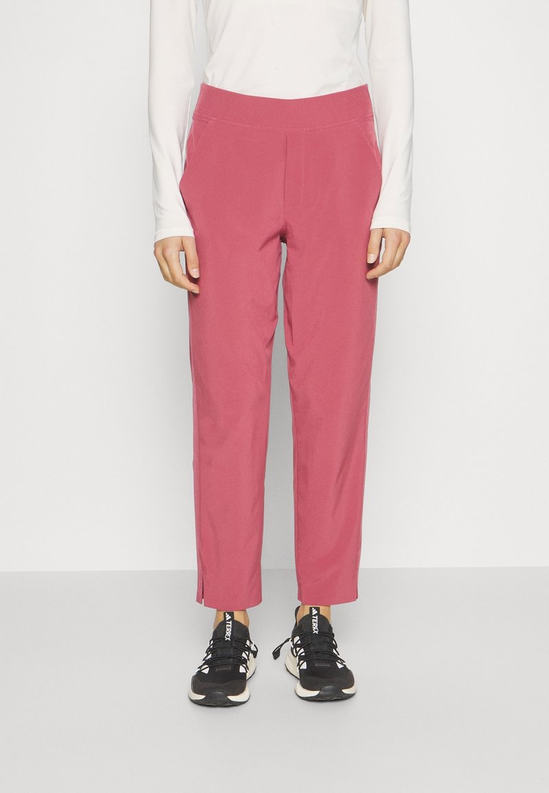 Personne portant un pantalon rose taille haute, coupe droite, avec des poches latérales et des baskets noires, debout devant un fond uni.