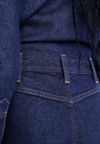 Jeans din denim albastru închis cu cusături portocalii, talie înaltă, buzunare pe spate și o textură netedă cu un design structurat.