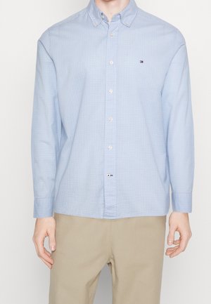 Homme portant une chemise bleu clair à petits carreaux avec boutons et un pantalon beige, debout devant un fond uni.