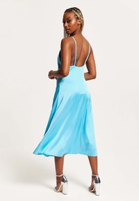 A sleeveless, backless dress in bright blue satin fabric with a flowing skirt. Silver heeled sandals complement the outfit.