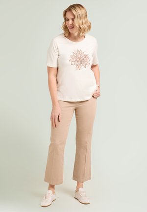 Kobieta w beżowych spodniach capri i białej koszulce z kwiatowym nadrukiem, stojąca z jedną ręką w kieszeni, beżowe trampki.