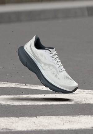 Scarpa da corsa bianca e grigia con lacci, che sembra fluttuare sopra una strada asfaltata con segnaletica bianca e piccoli detriti sparsi intorno.