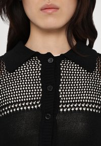 Black knitted cardigan featuring a mesh design with a solid collar, five buttons, and ribbed details at the waist and neckline.