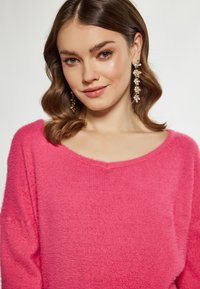 Maglione rosa, morbido, con scollo ampio e maniche lunghe, abbinato a orecchini d'oro decorati con piccole pietre preziose.