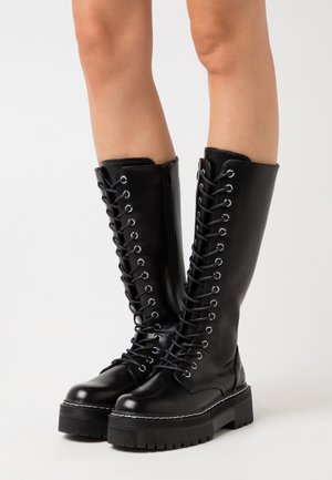 Stivali neri in pelle con lacci, alti fino al ginocchio, con occhielli in argento, suola piatta nera spessa e cerniera laterale per una facile vestibilità.