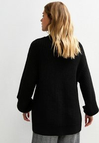 Maglione nero in maglia con texture, caratterizzato da una vestibilità ampia e maniche oversize, con orlo e polsini a costine. Indossato con pantaloni a fantasia.