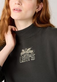Sötétzöld pulóver magas nyakkal, amelyen egy világosszínű, vintage stílusú Levi's logó és egy sas grafika található elöl.