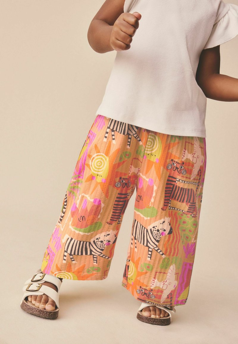 Niño con pantalones anchos de colores con estampados abstractos de animales, camiseta blanca y sandalias blancas con hebillas.