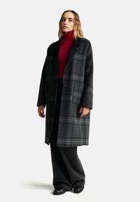 Abrigo de lana a cuadros en tonos grises, con diseño de doble botonadura, grandes bolsillos y cuello en pico, combinado con un suéter de cuello alto rojo y pantalones negros.