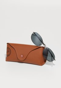 Ray-Ban aviacijos akiniai su tamsiais lęšiais gulintys ant rudo tekstūruoto odinio dėklo ant balto fono.