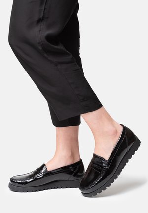Black patent leather slip-on shoes with a low, textured sole and stitched accents, worn with cropped black pants.