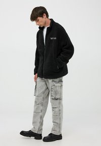 Czarna polarowa kurtka z zamkiem z przodu i wysokim kołnierzem; posiada haftowany napis. W zestawie z jasnoszarymi spodniami cargo i czarnymi butami.