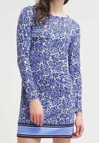 Långärmad klänning i blå blommönster med vita detaljer, med en enfärgad blå fåll och marinrandiga detaljer. Tyget verkar slätt och lätt.