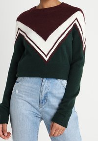 Donna che indossa un maglione verde scuro con strisce a zigzag bianche e bordeaux, abbinato a jeans chiari a vita alta.