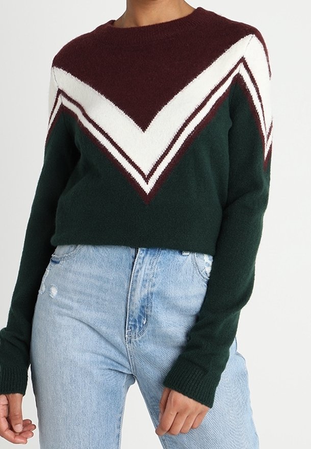 Donna che indossa un maglione verde scuro con strisce a zigzag bianche e bordeaux, abbinato a jeans chiari a vita alta.