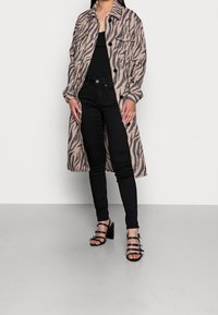Abrigo largo beige con rayas de tigre negras, con botones en la parte delantera, dos bolsillos en el pecho, usado sobre una blusa negra y jeans ajustados negros, combinado con sandalias de tacón negras.