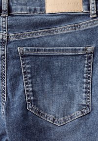 Jeans de mezclilla azul con un acabado ligeramente descolorido, que presentan un único bolsillo trasero. La costura es visible en un tono marrón claro, con una etiqueta de cuero en la parte superior.