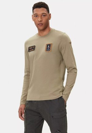 T-shirt a maniche lunghe di colore verde oliva in cotone. Presenta due toppe ricamate sul petto e un discreto scollo rotondo.
