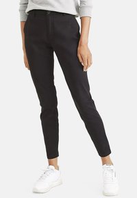 Čierne slim-fit chinos nohavice vyrobené z hladkej tkaniny, s gombíkovým zapínaním, plochým predným dielom a dĺžkou po členky, skombinované s bielymi teniskami.
