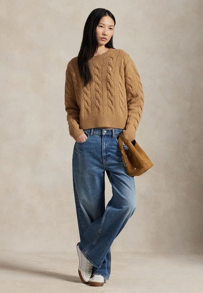 Mladá žena oblečená v béžovém svetr s copánkovým vzorem, volné modré džíny, bílé tenisky a držící hnědou semišovou kabelku, stojící na neutrálním pozadí.