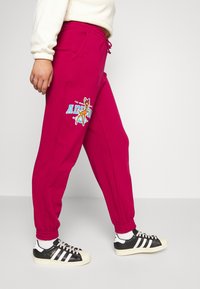 Person som bär ljusrosa träningsbyxor med en älggrafik och vit dragsko, i kombination med svartvita sneakers och vita strumpor.