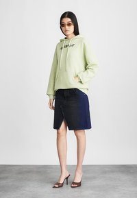 Zielona bluza z kapturem z przednią kieszenią, ściągaczami i kwiatową haftem, zestawiona z ciemną spódnicą jeansową z postrzępionym dołem i częściowym niebieskim fragmentem.