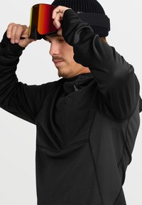 Veste en tissu noir avec une finition texturée et un col haut. Une personne ajuste des lunettes de ski teintées en rouge, portant un bonnet en tricot noir.