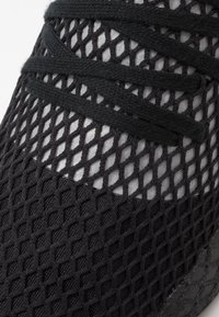 Svart träningssko med en övre del i mesh med diamantmönster, kompletterad med svarta platta snören och en solid svart gummisula.