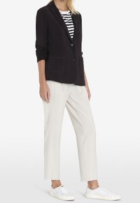 Blazer nero con due tasche anteriori, abbinato a una camicia a righe, pantaloni beige e sneakers bianche. Tessuto morbido, vestibilità su misura.