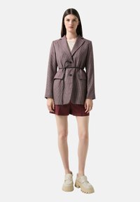 Kostkovaný blazer v burgundské a šedé látce, s klopovým límcem, dvěma předními kapsami a černým páskem na zavázání; stylizovaný s burgundskými šortkami a masivními botami.