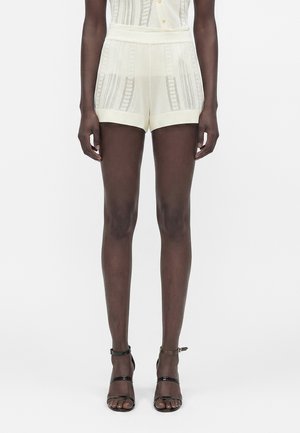 Γυναίκα που φορά ημιδιάφανα κρεμ σορτς με κάθετη ριγέ μπορντούρα και μαύρα πέδιλα με ανοιχτή μύτη και λουράκι στον αστράγαλο, στέκεται μπροστά σε λευκό φόντο.