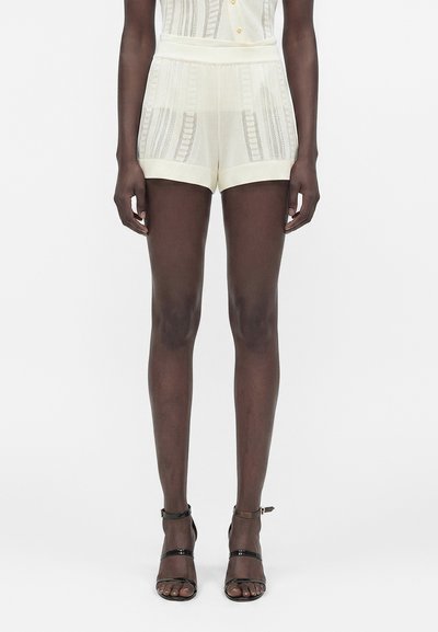Mujer con pantalones cortos crema semitransparentes con patrón de rayas verticales y tacones negros con tira alrededor del tobillo y punta abierta, de pie frente a un fondo blanco liso.