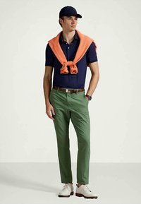 Camisa polo azul-marinho, suéter leve laranja drapeado, calças verdes, sapatos casuais e um boné azul-marinho, exibindo um estilo casual clássico.