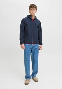 Marinejacke mit Kapuze, vorderem Reißverschluss und kontrastierenden gestreiften Akzenten. Über einem roten Hemd getragen, kombiniert mit blauen Jeans und beigen Schuhen.