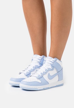 Jambes portant des baskets montantes Nike blanches et bleu clair avec des lacets, posées sur un fond blanc uni.